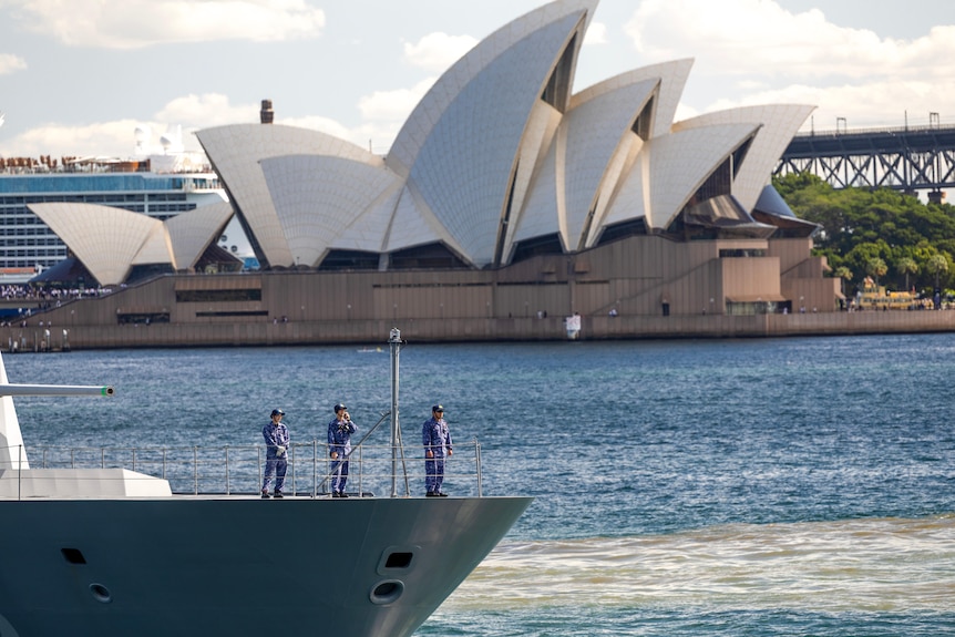 Three men in uniform stand on the deck of a grey frigate, which is setting off, with Sydney Opera House behind it.