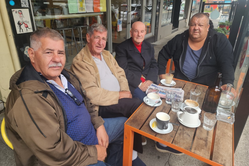 Four men look at the camera, as they sit around a cafe table on a footpath.