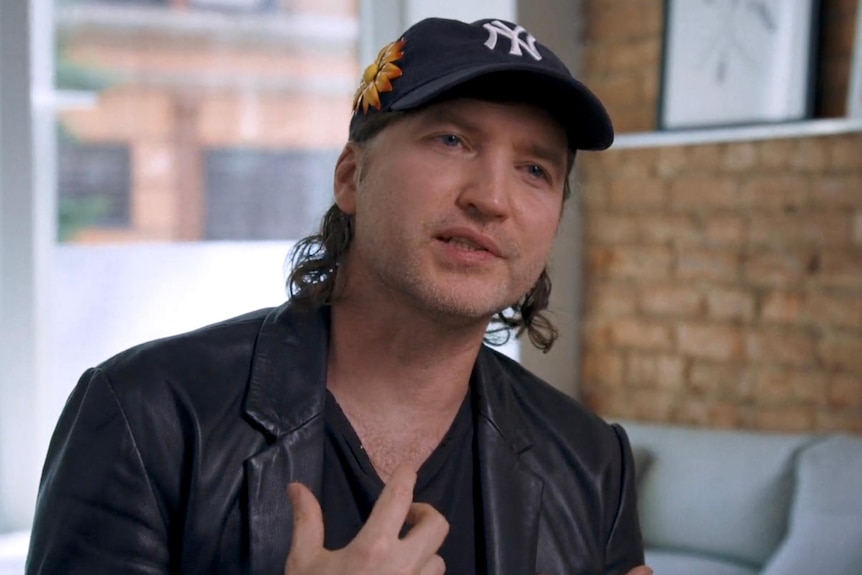 middle-aged man wears leather jacket, NYC baseball cap and gestures, sitting in an office