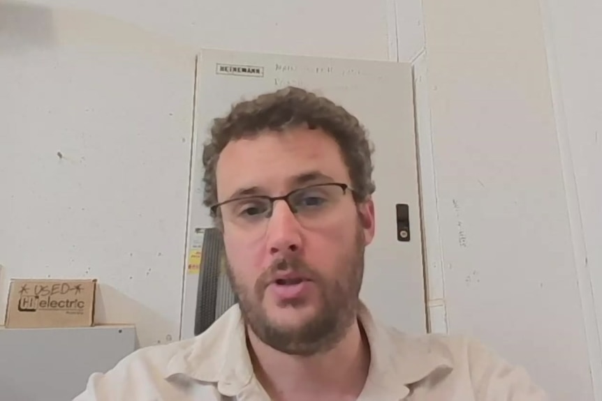Joe Calvert has curly hair and glasses and is sitting in a room with white walls and a door behind him.