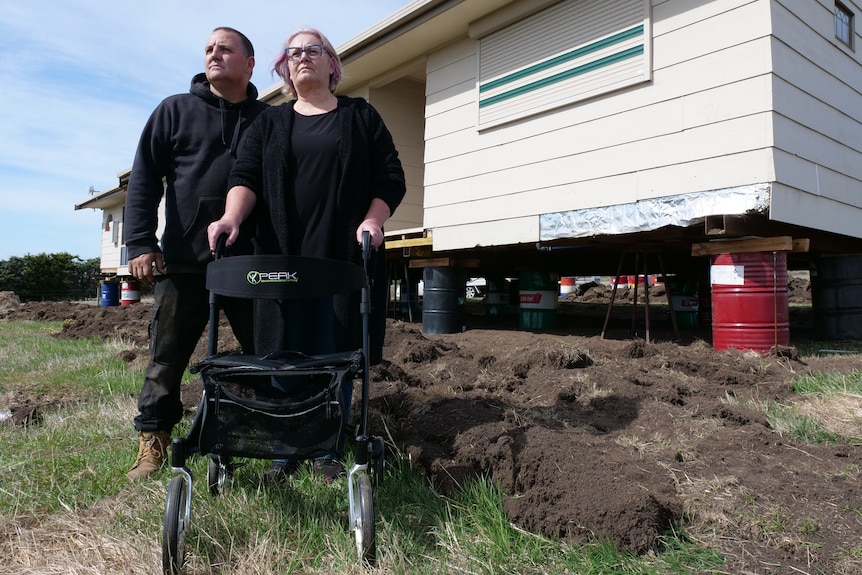 a man and a woman with a walker stand in front of a home in a paddock