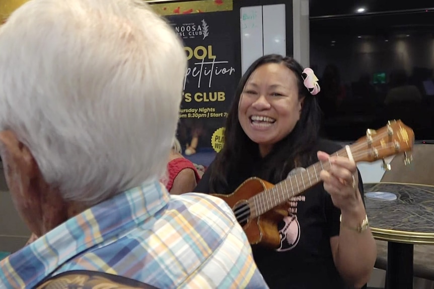 A smiling lady playing a wooden ukulele in an RSL club.