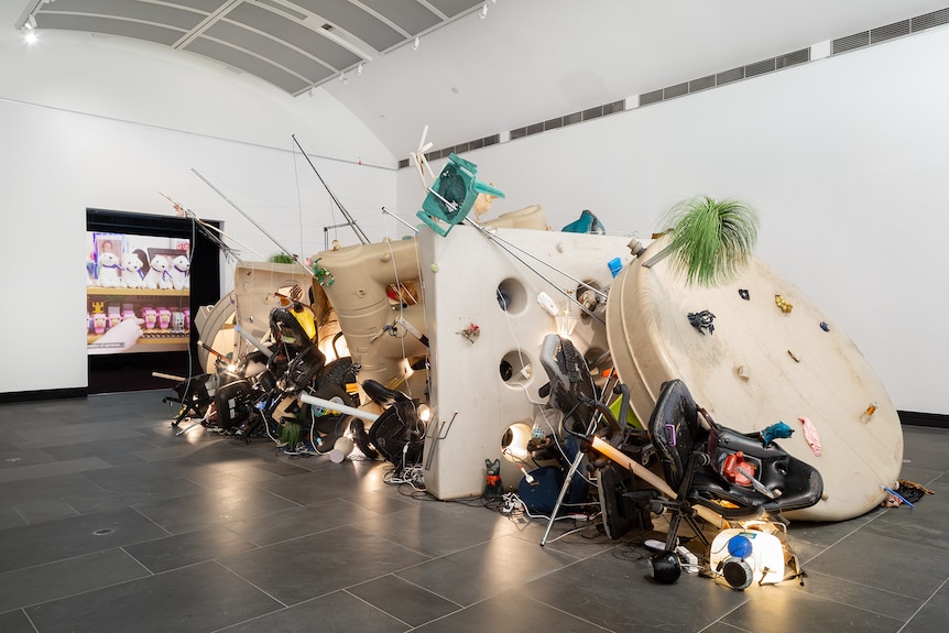 In a white gallery sits a pile of rubbish, mostly made of plastic. The piled rubbish includes computer chairs and lamps.