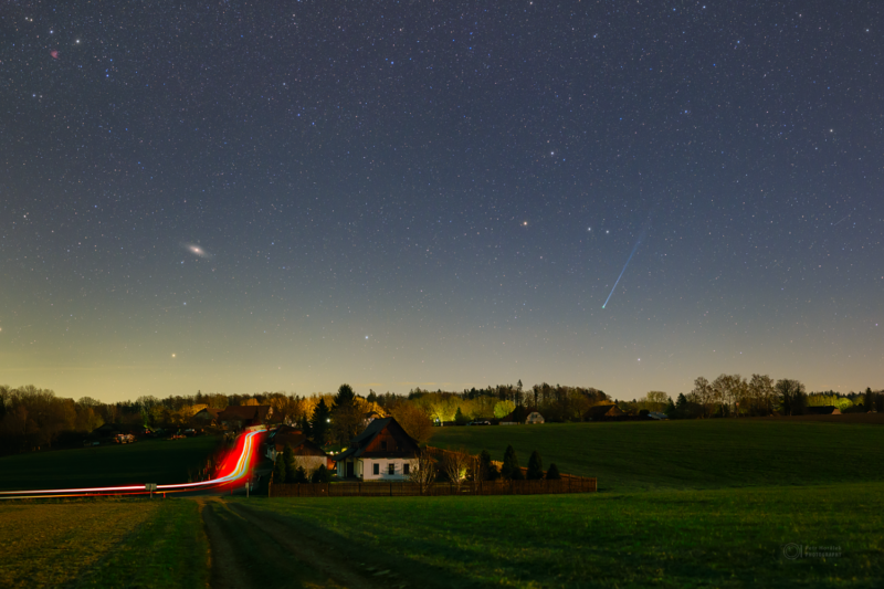Car lights streaking through the scenery with a comet at right in the sky and a fuzzy galaxy at left.