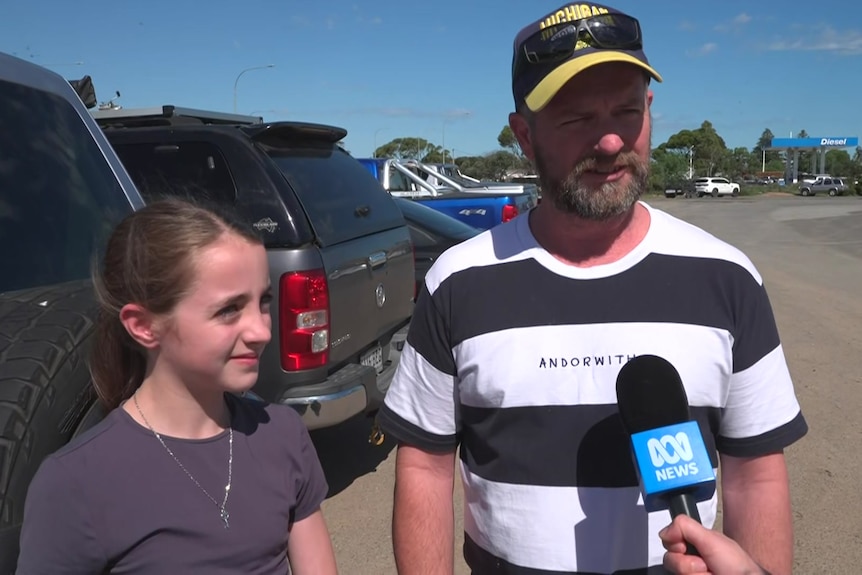 A young girl stands next to her dad with a row of cars behind them and a service station in the distance.