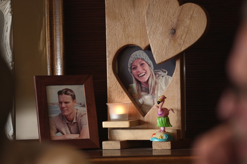 Two photos in wooden frames on a mantle, one of a middle-aged man, the other young blonde woman.  A candle sits between the two