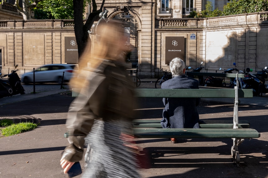A blurred woman walks past a park bench with someone sitting on it.