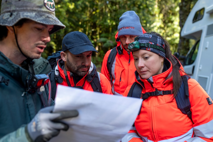 Two men and a woman, all wearing orange hi-vis clothing and head gear, listen as a man in grey and a bucket hat talks