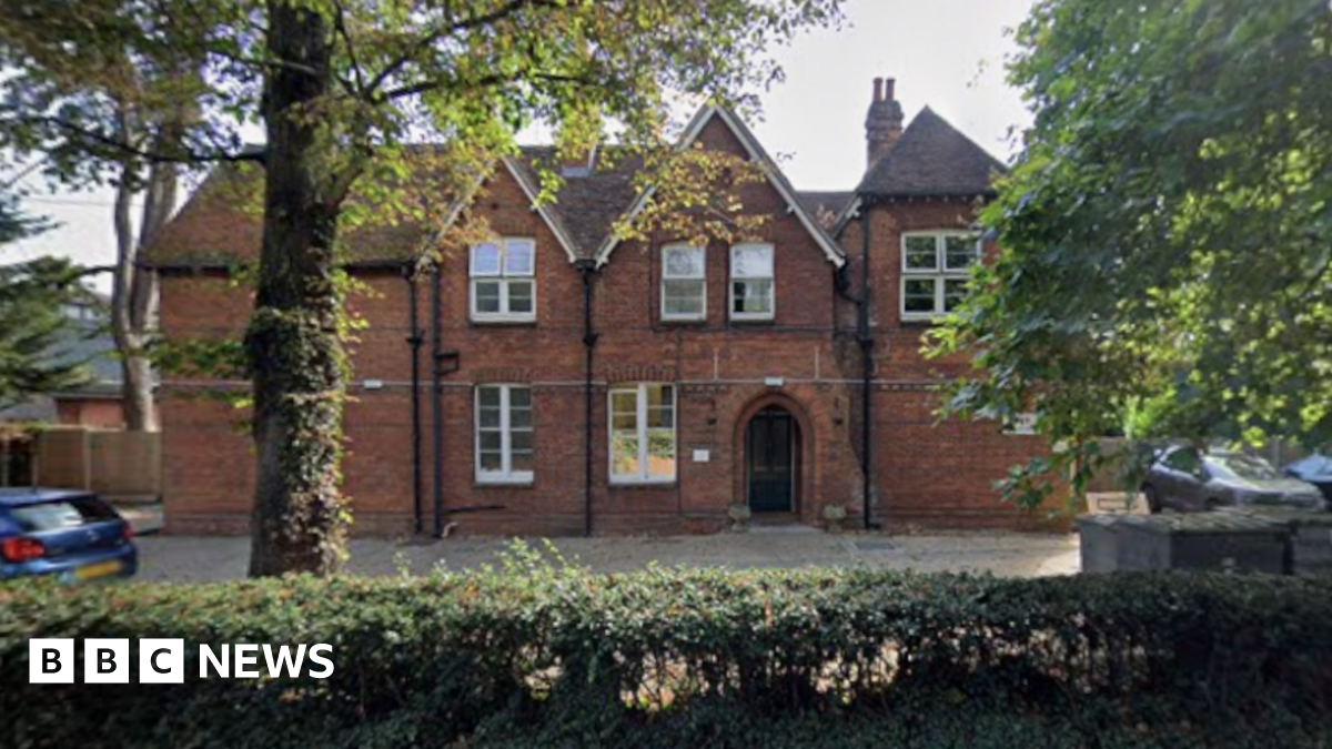 A red-brick building stands behind trees and bushes. The building has two apex roof sections and six large white-framed windows. It has a door arch for an entrance and a paved area out the front used as a car park.