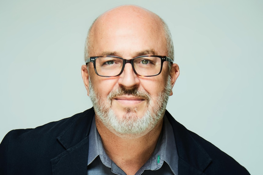 Man with grey facial hair and bald head in suit and wearing dark glasses
