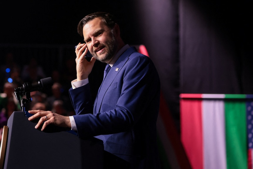 JD Vance smiles as he stands at a podium with a mobile phone to his ear.