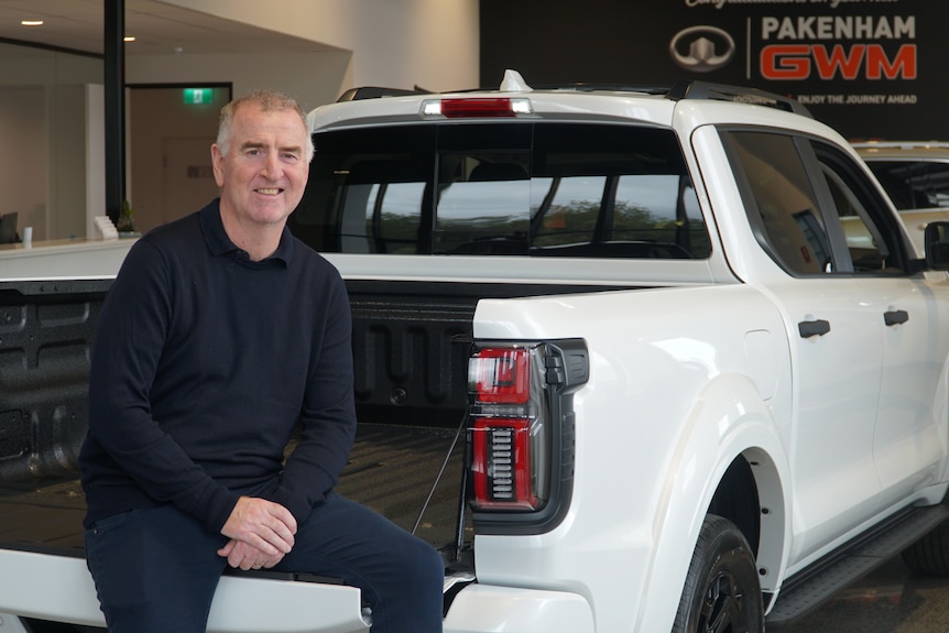 John sitting on a white ute, smiling, wears all black. Caucasian.