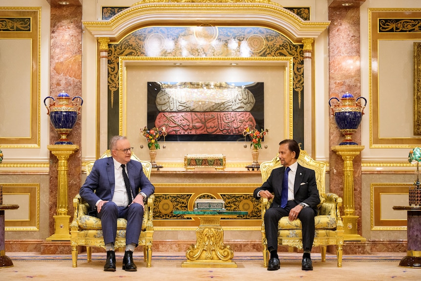 Two men wearing suits sit in gold chairs in a room full of gold and marble decorations.