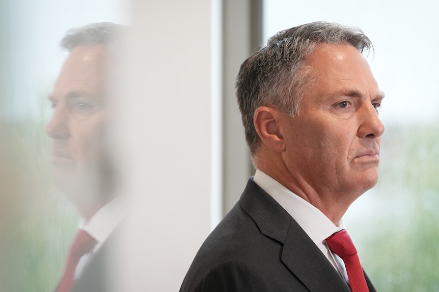 A man wearing a suit and red tie looks seriously to the side.