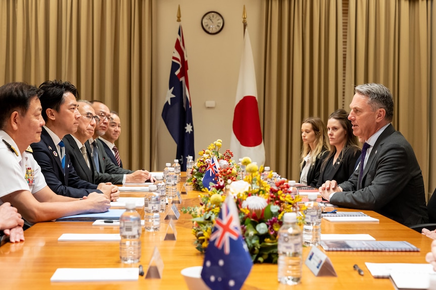 Koizumi Shinjiro and Richard Marles sit looking at each other at the opposite sides of a table alongside other people. 