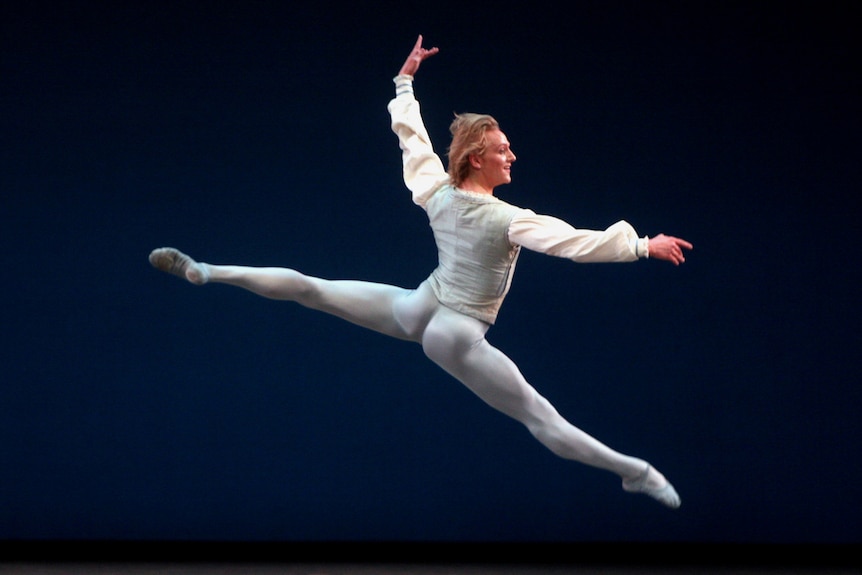 David Hallberg, in tights and a loose top, leaps with arms and legs wide across a dark stage