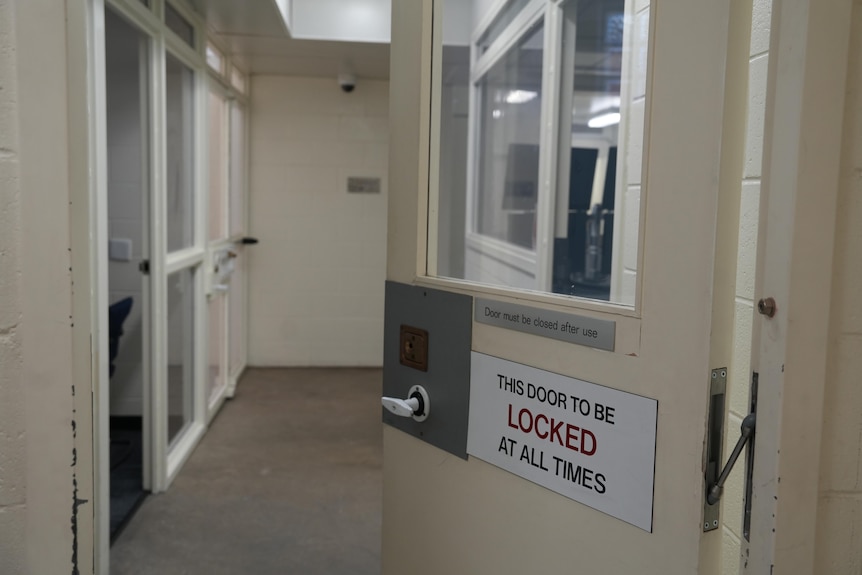 An opened door with a sign 'this door to be locked at all times', leads to a hallway with rooms and glass windows on both sides