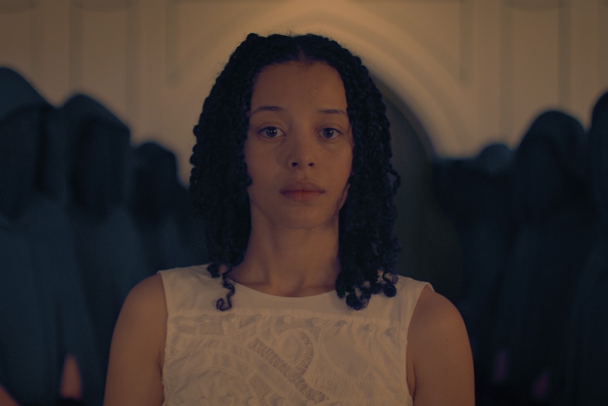 A young Black woman with natural hair surrounded by people in hoods with faces not visible