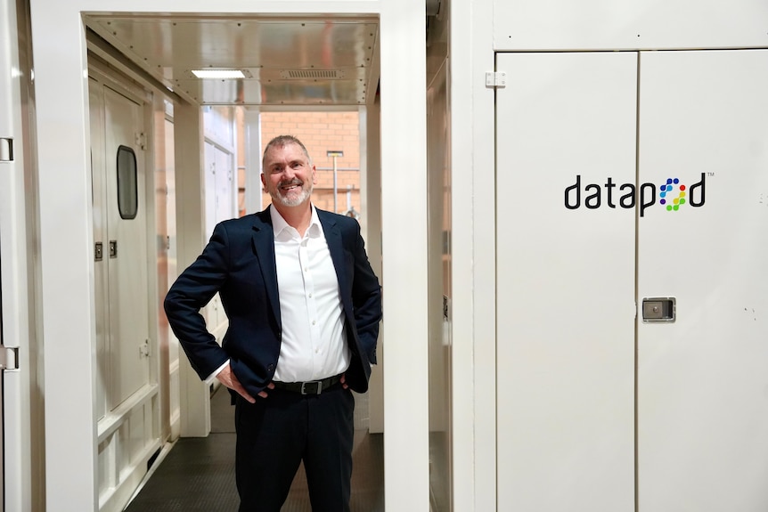 A man wearing a suit and white shirt stands smiling with hands on hips near a Datapod sign.