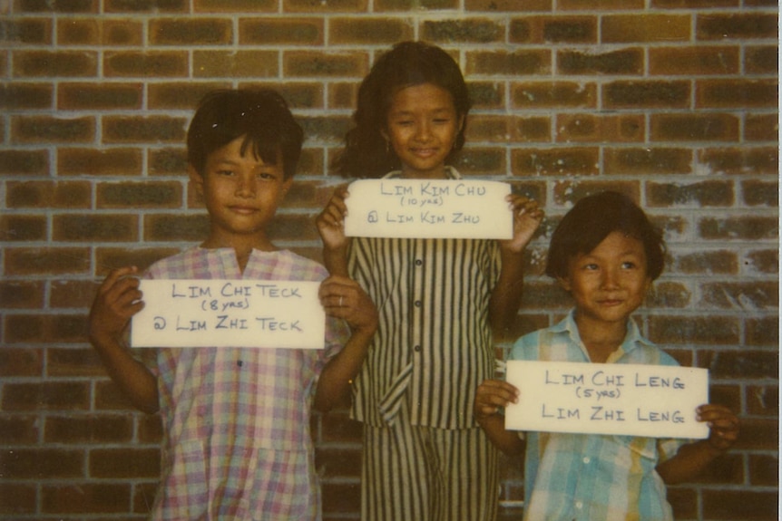 Kim and two of her brothers stand holding a sign with their names and ages..