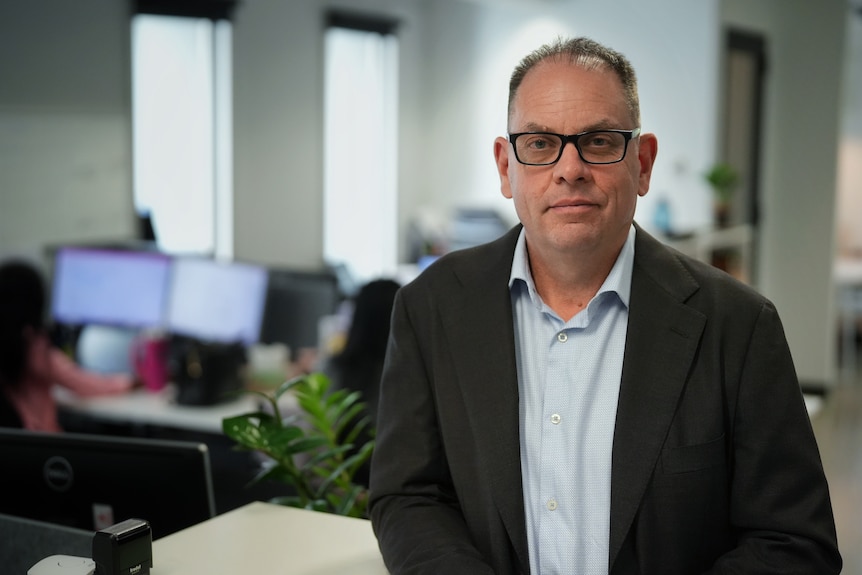 A man stands in an office leaning against a shelf, wearing a suit and glasses.