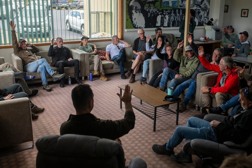 A large group of people, male and female, sit in a semi circle in chairs, their hands raised 