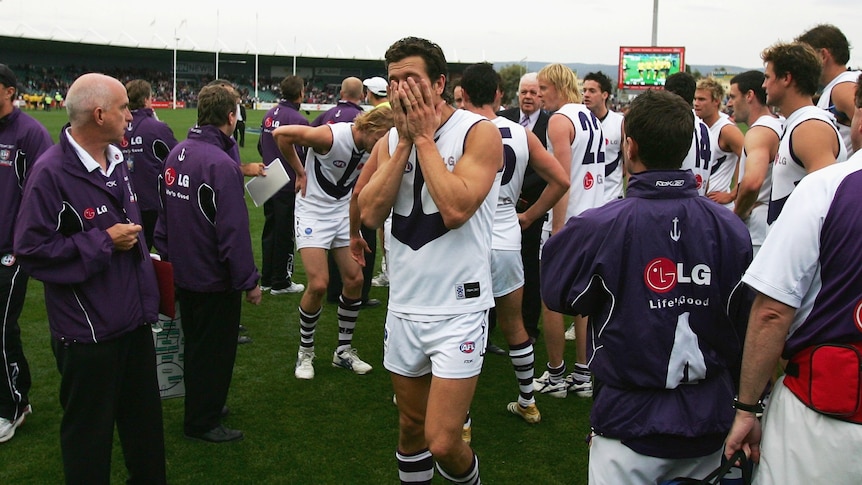 A Fremantle AFL player walks off the ground with his hands over his face, followed by teammates after a game.