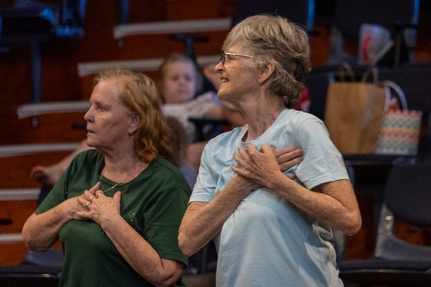Two women aged in their sixties hold their hands on their hearts; one wearing green the other blue with glasses.