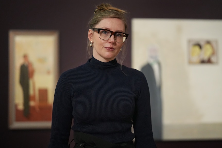 A woman with blonde hair and glasses stands in an art gallery smiling lightly.