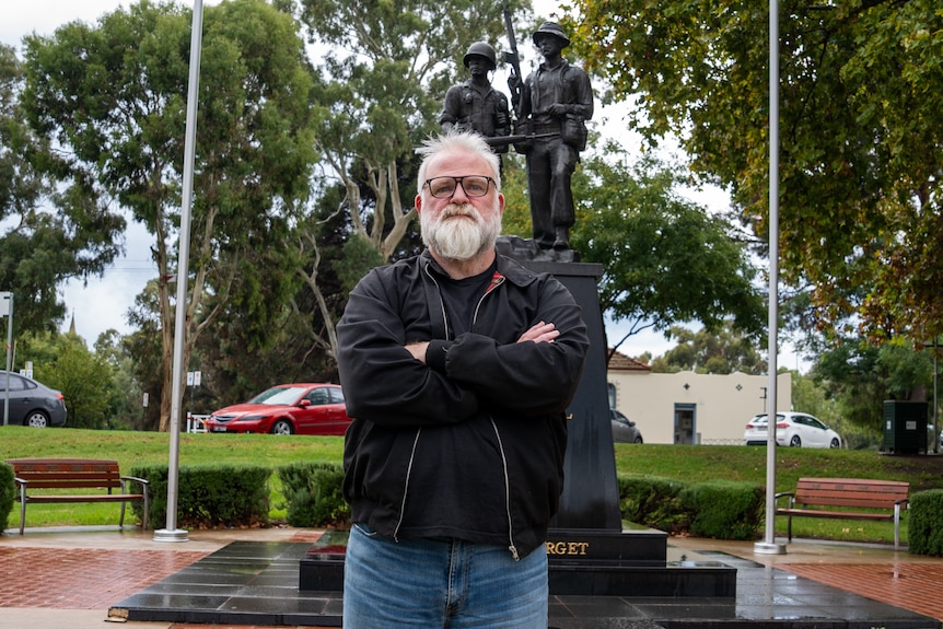 A man with white hair and a beard stands with his arms crossed in front of a war memorial