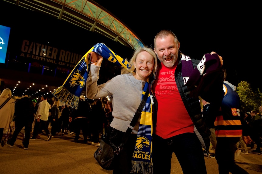 A woman with blonde hair and a bald man have their arms around each other holding up their footy scarves cheering. 