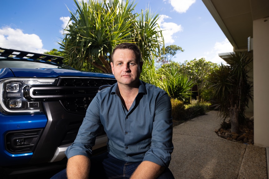 A man in a blue shirt sitting in front of a blue ute in his driveway.
