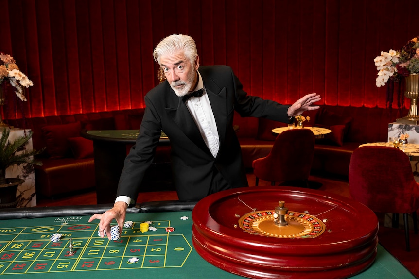 Shaun leans over a green gambling table in a luxe looking casino room while wearing a tuxe, with his hair swept back.