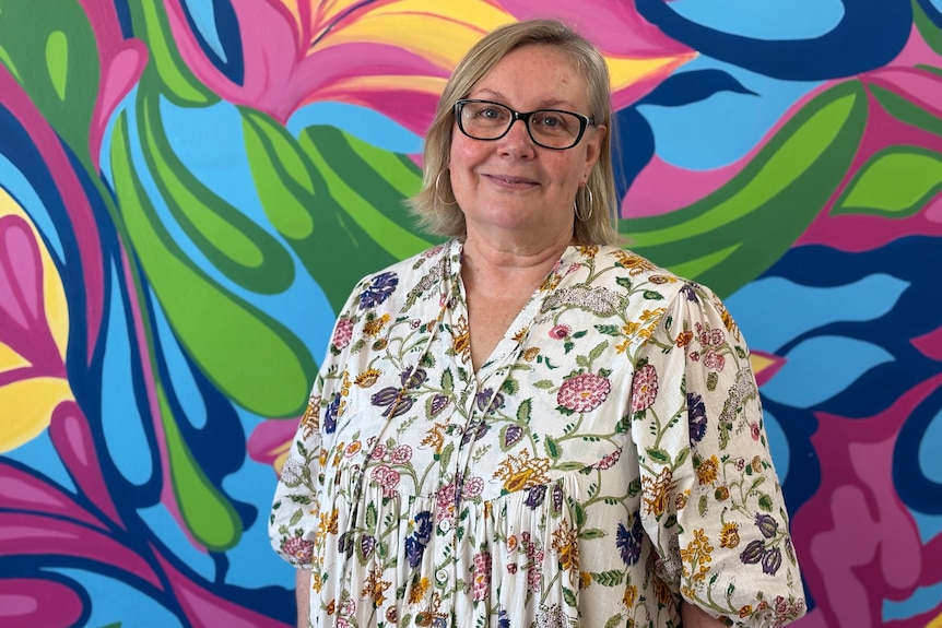 A woman with dark-framed glasses and short hair wearing a bright flowery dress stands in front of a colourful mural.