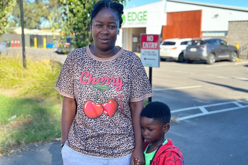 A woman in a colourful t-shirt holds her son's hand, who is shy, at a car park outside a Edge early learning centre