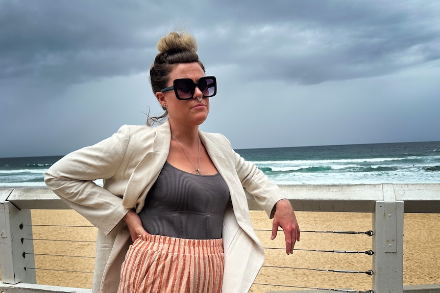 A woman with her hair in a bun wears sunglasses and an open blazer while leaning against a railing at a beach.