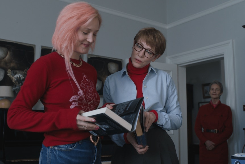 Two women stand next to each other in a room, a third woman stands in the doorway