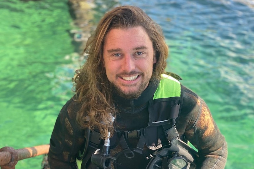 Stefan in a surfer wetsuit carrying a large camera, light equipment and flippers while walking up step from the ocean.