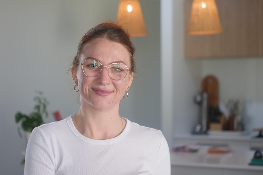 A smiling, dark-haired woman in glasses sits in a kitchen area.