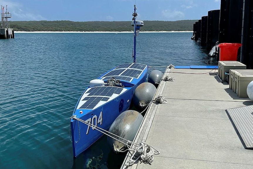 A blue vessels that looks like an enclosed canoe with solar panels on top sits on the water. 
