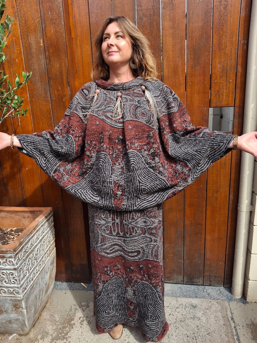 Middle aged woman wearing a knitted skirt and poncho in russet red and grey colours. her arms are spread out wide