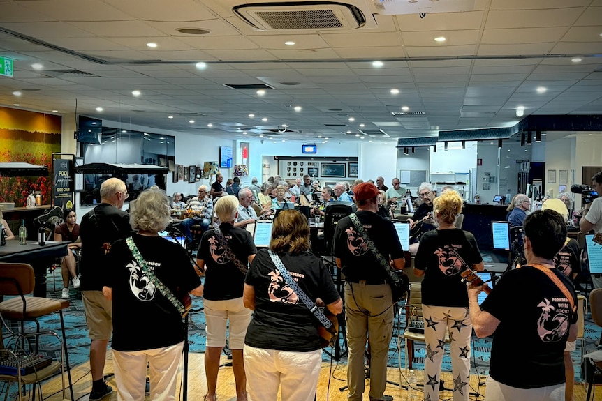 Seven people stand on stage playing Ukuleles, in front of a crowd of sitting ukulele players.