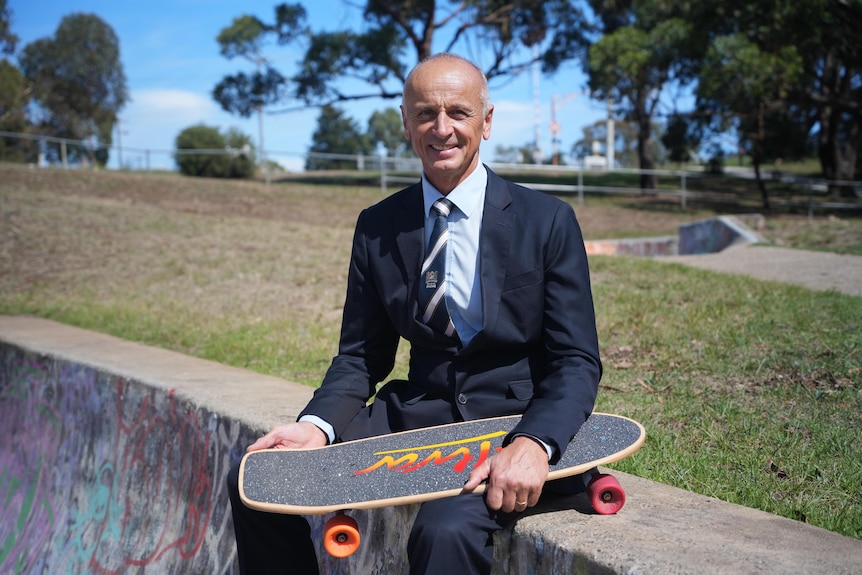 A man wearing a suit holds a skateboard on his lap.