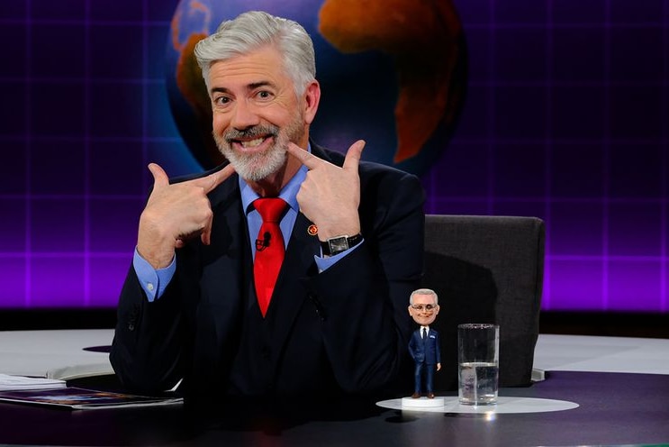 A grinning Shaun Micallef sitting behind a news desk