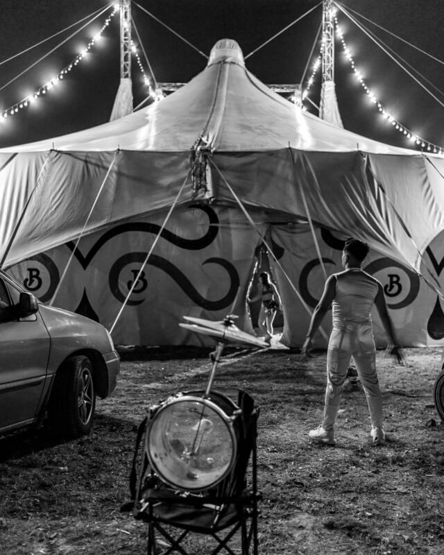 A black and white photo of a circus tent at night. A performer in costume stands in front, facing an acrobat hanging from a rope at the entrance. Drums and a parked car are visible in the foreground.