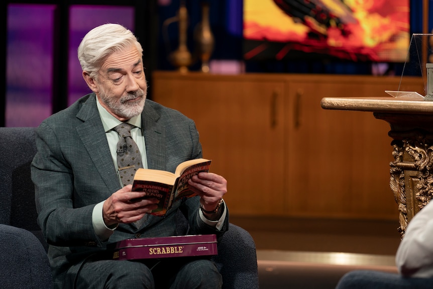 A TV still of Shaun Micallef, early 60s, looking intently at a book on a TV set. He has a Scrabble box on his lap.