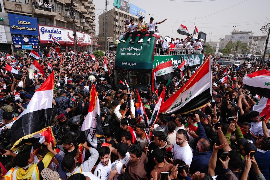 Members of Iraq's men's football team are on an open top bus, surrounded by fans waving Iraqi flags