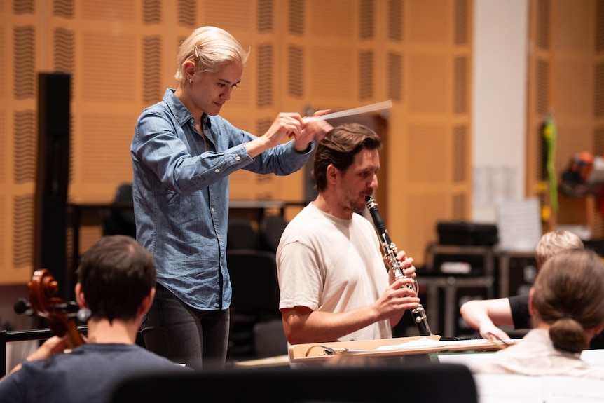 A woman with short, blonde hair waves a conductor's baton as she stands behind a clarinettist who is facing an orchestra.