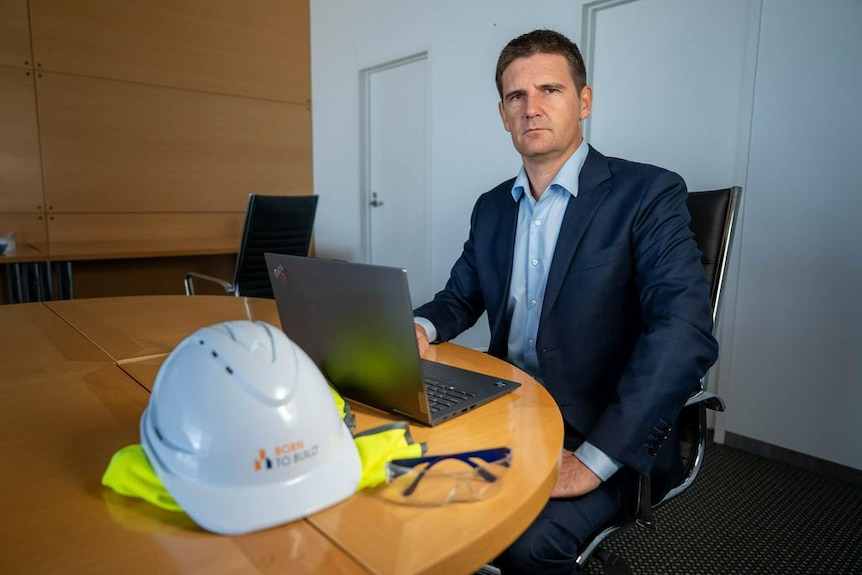 A dark-haired man in a dark suit sits at a laptop near a hard hat and high-vis vest on a desk in an office.