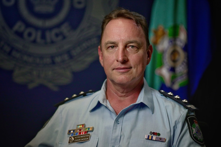 A portrait of a man in a Queensland police uniform with the Queensland police logo on the wall in the background and a flag.